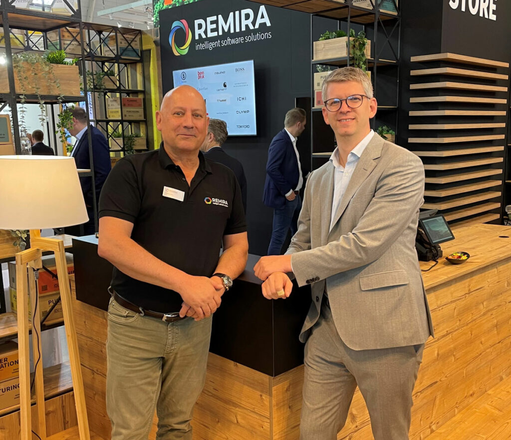 Zwei Männer, einer im schwarzen Poloshirt, der andere im grauen Anzug, stehen lächelnd vor einem REMIRA-Messestand bei einer Veranstaltung. Im Hintergrund sind weitere Teilnehmer und ein Bildschirm mit verschiedenen Logos zum Thema Konditionenmanagement zu sehen.