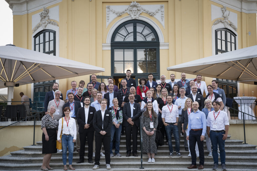 A group of people, some wearing conference badges and lanyards, pose for a photo on the stairs of a yellow building with a large arched window. They appear to be delegates finalizing details for the Jahresvereinbarung.